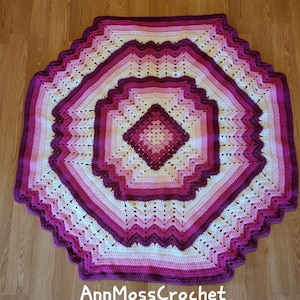 May include: A crocheted blanket in shades of pink and white with a geometric pattern. The blanket is made in a hexagon shape with a smaller diamond shape in the center. The text "AnnMossCrochet" is visible in the bottom center of the image.
