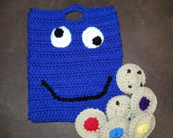 Op de afbeelding: Blauwe gehaakte tas met een glimlachend gezicht en oogjes. De tas is gevuld met kleurrijke gehaakte koekjes.