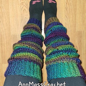 Könnte beinhalten: Handgefertigte Häkel-Beinstulpen in Blau-, Grün-, Lila- und Brauntönen. Die Beinstulpen werden über schwarzen Socken getragen. Der Text "AnnMossCrochet" ist unten im Bild zu sehen.