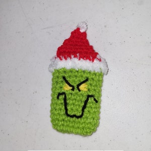 May include: Green crocheted Grinch character wearing a red and white Santa hat. The Grinch has yellow eyes and a black mouth. The image includes the text "crochet pattern".