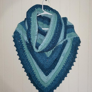 May include: A crocheted triangular shawl in shades of teal and blue, hanging on a white hanger against a white wood panel background.