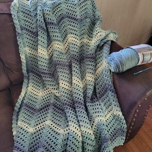 Puede incluir: Una manta de ganchillo hecha a mano en tonos de azul, verde y blanco, sobre una silla de cuero marrón. La manta presenta un patrón de chevrón y un diseño texturizado. Se ve un ovillo de hilo a juego y un ganchillo.