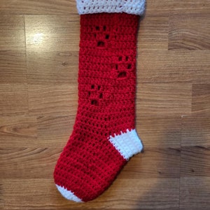 Puede incluir: Calcetín de Navidad de crochet rojo y blanco con un puño y una punta blancos. El calcetín tiene un diseño de copo de nieve.