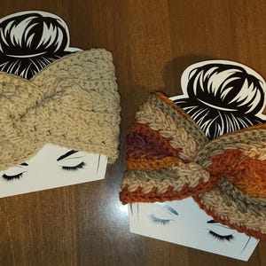 May include: Two handmade cable knit headbands. One is a solid beige color, and the other is a mix of orange, brown, and beige. Both headbands are displayed on white cards with a black and white illustration of a woman's face.