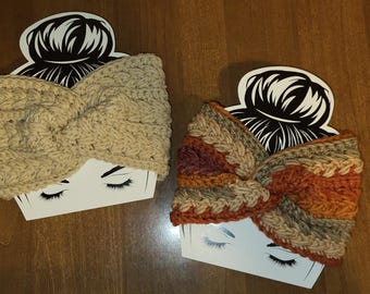 Op de afbeelding: Twee handgemaakte kabelgebreide hoofdbanden. Eén is effen beige van kleur, de andere is een mix van oranje, bruin en beige. Beide hoofdbanden worden gepresenteerd op witte kaarten met een zwart-wit illustratie van een vrouwengezicht.