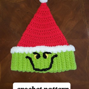 May include: A handmade crochet hat in the style of a Christmas character. The hat features a red pointed top with a white pom-pom, a green face with black and yellow details, and the text "crochet pattern".