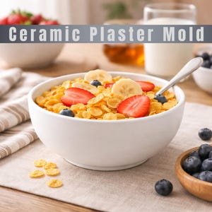 Könnte beinhalten: Eine weiße Keramikschüssel gefüllt mit Müsli, geschnittenen Erdbeeren, Blaubeeren und Bananenscheiben. Der Text "Ceramic Plaster Mold" wird über der Schüssel angezeigt. Ein Löffel liegt im Müsli. Rechts ist eine Holzschüssel mit Blaubeeren.