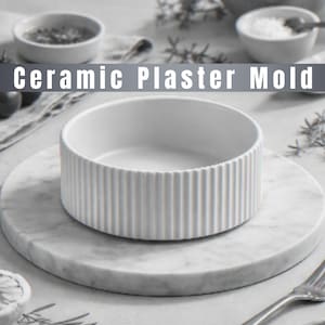 May include: A white ceramic bowl with vertical ribbed detailing, on a round marble platter. The text "Ceramic Plaster Mold" is above the bowl. Other small bowls and a fork are in the background.