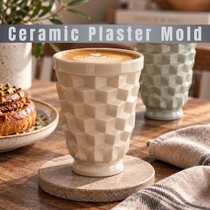 Puede incluir: Una taza de cerámica de color crema con un exterior de patrón de cubos geométricos, llena de café con arte latte. El texto "Ceramic Plaster Mold" aparece en la parte superior. La taza descansa sobre un posavasos redondo de piedra moteada.