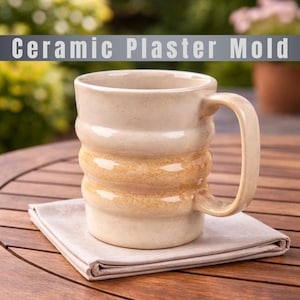 Może przedstawiać: Beżowy ceramiczny kubek z żebrowanym wzorem i zakrzywionym uchwytem. Kubek stoi na złożonej beżowej serwetce na drewnianym stole. Tekst "Ceramic Plaster Mold" znajduje się nad kubkiem.