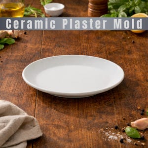 Può includere: Un piatto in ceramica bianco con un bordo leggermente rialzato. Il piatto è centrato su una superficie di legno scuro. Il testo "Ceramic Plaster Mold" è visualizzato sopra il piatto in un banner grigio.