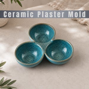 Pode incluir: Um molde de gesso cerâmico de três seções com um esmalte salpicado vibrante em azul e verde-azulado. O prato consiste em três tigelas circulares interconectadas com bordas lisas e arredondadas e um acabamento brilhante em todas as superfícies.