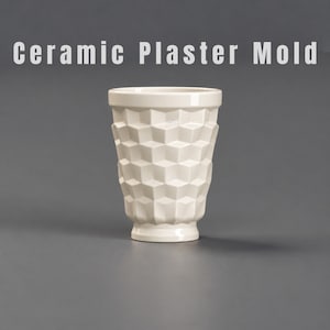 Könnte beinhalten: Eine cremefarbene Keramik-Gipsform für einen Becher mit einem sich wiederholenden geometrischen Würfelmuster auf der Außenfläche. Der Text Ceramic Plaster Mold ist in weißen Buchstaben über dem Objekt vor grauem Hintergrund zu sehen.