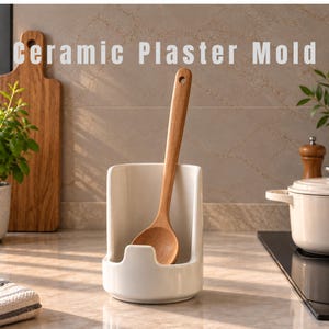 May include: A white ceramic spoon rest with a tall, curved back and a central notch, holding a long-handled wooden spoon. The text "Ceramic Plaster Mold" is visible at the top of the image. The spoon rest has a smooth, glossy finish.