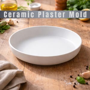 Pode incluir: Um molde de gesso cerâmico branco de formato redondo, com acabamento mate liso e uma base larga e rasa. O objeto apresenta um design limpo com fundo plano e bordas ligeiramente curvas e verticais para projetos de fundição ou artesanato.