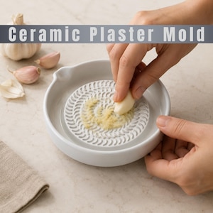 Puede incluir: Un plato rallador de ajo de cerámica blanca con un centro circular texturizado para moler dientes de ajo. El plato tiene un pequeño pico en el borde y se acompaña de dientes de ajo enteros. El texto Ceramic Plaster Mold es visible.