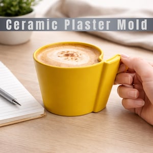 Könnte beinhalten: Eine gelbe Keramik-Kaffeetasse mit einem einzigartigen Griff, gefüllt mit Cappuccino. Die Tasse steht auf einer Holzoberfläche, neben einem Notizblock und einem Stift. Der Text "Ceramic Plaster Mold" befindet sich über der Tasse.