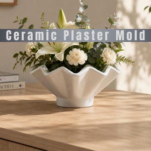 Puede incluir: Un cuenco de molde de yeso cerámico blanco con un diseño de borde festoneado y acanalado. El cuenco está lleno de un arreglo floral con lirios blancos, claveles blancos y hojas de eucalipto verdes. El texto Ceramic Plaster Mold es visible.