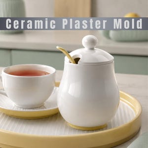 Könnte beinhalten: Eine weiße Zuckerdose aus Keramik mit passendem Deckel und einem kleinen gelben Löffel, begleitet von einer weißen Teetasse auf einer Untertasse, alles auf einem runden gelben Tablett. Der Text Ceramic Plaster Mold ist oben zu sehen.