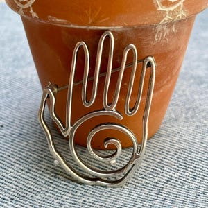 May include: A silver-toned metal hand-shaped brooch with a spiral design in the palm. The brooch is set against a terracotta pot with bird drawings. The background is a blue denim fabric.