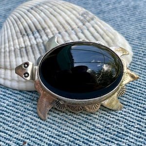 May include: A silver-toned turtle-shaped brooch with a large, oval, black stone center. The brooch has detailed flipper and head designs. The background includes a white seashell and a blue denim surface.