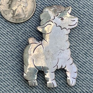 May include: A silver-toned metal sheep brooch. The brooch is in the shape of a sheep with detailed features. A U.S. quarter coin is next to the brooch for size comparison. The brooch is on a blue denim background.