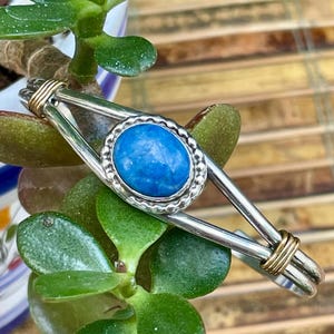 May include: A silver-tone bracelet with a vibrant blue oval stone at its center. The stone is set in a textured silver bezel. The bracelet features gold-tone wire accents and a unique open design, resting on green foliage.