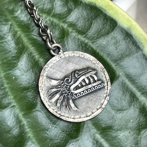 May include: Silver pendant necklace featuring a detailed, stylized silver design of a mythical creature's head with a feathered headdress.
