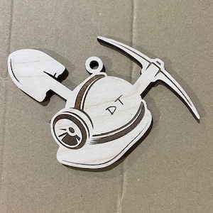 May include: A wooden ornament featuring a mining helmet with a pickaxe and shovel. The helmet has a lamp and the initials "DT" etched on it. The ornament is made of light-colored wood.