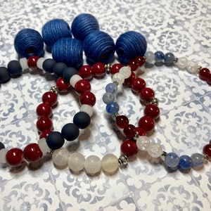 May include: Three bracelets made with red, white, and blue beads. The bracelets are made with different types of beads, including matte black, white agate, and blue agate.
