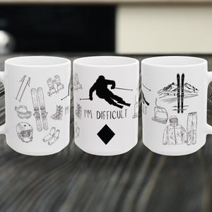 May include: White ceramic mugs with black line art illustrations of skiing equipment and a skier. One mug has the text "I'M DIFFICULT" with a black diamond symbol.