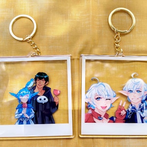 Puede incluir: Dos llaveros de acrílico transparente con anillos dorados. Cada llavero presenta una colorida ilustración de estilo anime. Uno muestra dos figuras, y el otro dos figuras diferentes. Los llaveros son cuadrados y tienen un borde blanco.