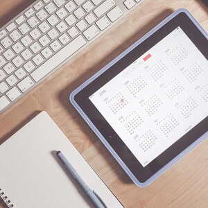 Pode incluir: Vista aérea de um espaço de trabalho com um tablet exibindo um calendário, um teclado, um smartphone, um bloco de notas e uma caneta. O tablet está no centro, com o teclado e o smartphone à esquerda. O bloco de notas e a caneta estão no canto inferior esquerdo.