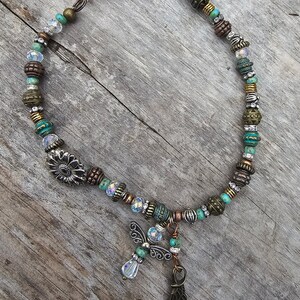 May include: A turquoise and silver beaded necklace with a sunflower charm and an angel wing pendant. The necklace is made with a variety of beads, including turquoise, silver, and copper.
