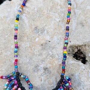 May include: A colorful beaded necklace with a fringe of beads at the bottom. The necklace is made with a variety of colors, including blue, green, purple, pink, yellow, and orange. The necklace is on a light gray background.