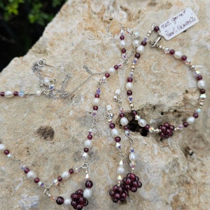 May include: A set of pearl and garnet jewelry, including a necklace and earrings. The necklace features alternating white pearls and small, dark red garnet beads. The earrings are drop earrings with a cluster of garnet beads at the bottom.
