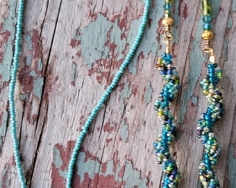 May include: A beaded eyeglass chain and a matching beaded necklace are displayed on a weathered wooden surface. The eyeglass chain features a turquoise beaded strand and two beaded spiral chains in shades of blue, green, and gold. The necklace is a simple turquoise beaded strand.