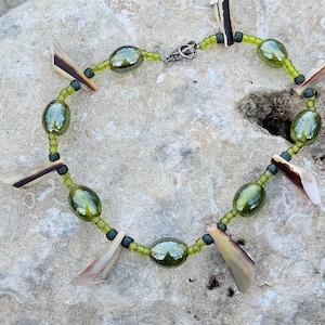 May include: A green glass bead necklace with shell accents. The necklace is made with small green beads and larger oval-shaped green glass beads. The shell accents are a light brown color and are shaped like triangles.