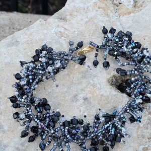 May include: A beaded bracelet featuring a mix of black, gray, and iridescent beads. The bracelet has a gold-colored clasp and is made with various shapes and sizes of beads, creating a textured and detailed design. The bracelet is displayed on a light-colored stone surface.