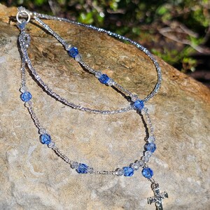 May include: A silver chain necklace with a silver cross charm. The necklace is made with small clear and blue beads.