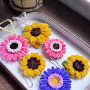 Puede incluir: Cinco llaveros de flores de fieltro de colores vivos en una caja blanca. Las flores son amarillas, rosas, moradas y marrones. Los llaveros están unidos a cadenas plateadas.