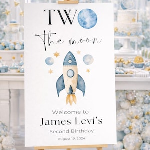 May include: A white sign on a wooden easel with the text "TWO the moon" and a watercolor rocket ship illustration. The sign welcomes guests to James Levi's second birthday on August 19, 2024.