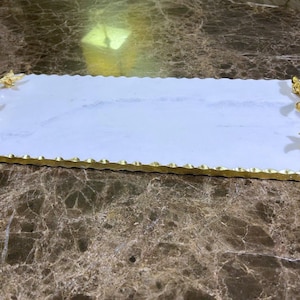 Op de afbeelding: Wit marmeren dienblad met bladgouden accenten en een decoratief, sierlijk, bladvormig handvat aan elke kant.