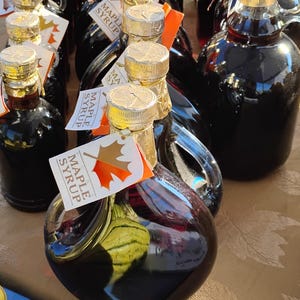 May include: Bottles of maple syrup with gold foil-covered lids and attached tags. The tags read "Maple Syrup" and feature a maple leaf. The syrup is in clear glass bottles.