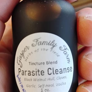 May include: A black glass bottle with a dropper top, held in a hand. The bottle has a white label with text that reads "Parasite Cleanse" and lists ingredients. The bottle contains 1 oz of liquid.