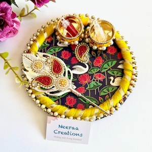 May include: A circular decorative tray featuring a dark background with a lotus flower and swan pattern. The rim is wrapped in yellow thread and gold ribbon with pearl accents. It holds two small gold containers and a silver feather-shaped ornament.
