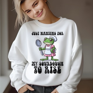 Puede incluir: Sudadera blanca con una rana verde que lleva un gorro de cocinero y un delantal sosteniendo una cuchara. El texto en la sudadera dice "Just wanting for my sourdough to rise".