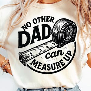 Puede incluir: Camiseta color crema con un gráfico negro que presenta una cinta métrica y el texto "NO OTHER DAD can MEASURE UP". La cinta métrica está detallada con números y marcas.