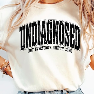 Könnte beinhalten: Cremefarbenes Sweatshirt mit dem Text "UNDIAGNOSED" in einer fetten, schwarzen, verwitterten Schrift, die sich über der Brust wölbt. Darunter steht der Satz "BUT EVERYONE'S PRETTY SURE" in kleinerer, schwarzer Schrift.