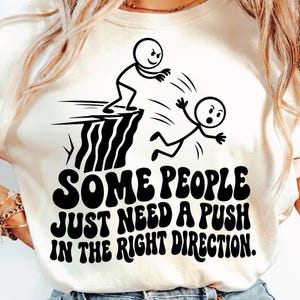 以下が含まれることがあります： クリーム色のTシャツ。黒い棒人間のグラフィック。一人が崖からもう一人を突き落とす。テキストには「SOME PEOPLE JUST NEED A PUSH IN THE RIGHT DIRECTION」と書かれている。